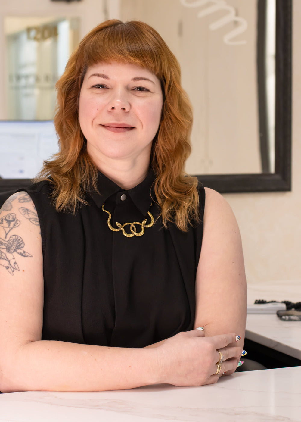A woman with red hair wearing a sleeveless black top and a gold necklace, sitting at a white table in an indoor setting.